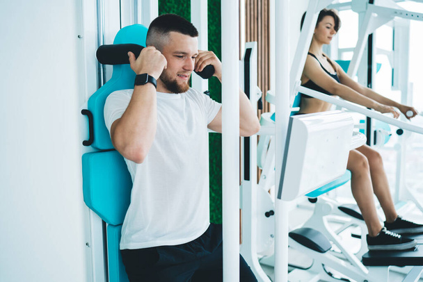 Deportista barbudo concentrado haciendo esfuerzo al hacer ejercicio en la máquina deportiva en el gimnasio durante el entrenamiento con la mujer y mirando hacia abajo - Foto, imagen