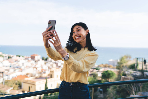 Asian female vlogger grinning while clicking selfie images via cellular front camera using roaming internet connection during travel vacations - Photo, Image