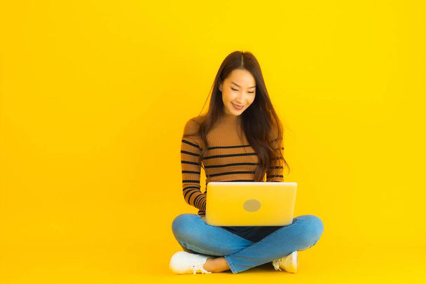 Portret mooie jonge aziatische vrouw zitten op de vloer voor gebruik laptop of computer op geel geïsoleerde achtergrond - Foto, afbeelding