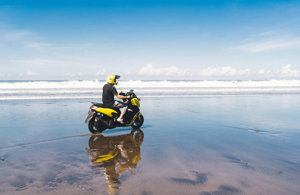 Side view of anonymous male traveler in stylish outfit and yellow helmet riding modern motorbike along wet sandy beach of waving ocean - Foto, immagini