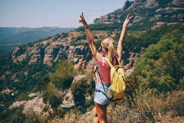 Back view of young female standing on hill putting hands up and showing v sign while hiking alone during summer holidays - Фото, изображение