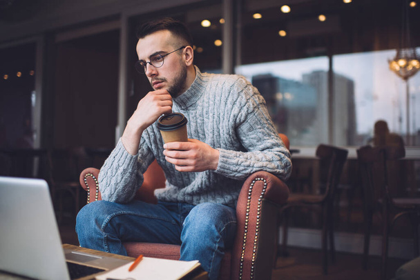 Concentrated male in eyeglasses with paper cup of hot drink looking at laptop while working remotely on new business project - Foto, Imagen