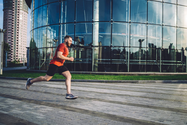 Side view of athletic sportsman in sport clothes running fast on pavement near modern building in city area in bright sunny day - Fotoğraf, Görsel