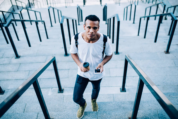 From above of smiling young African American man in casual wear and with backpack messaging on mobile phone while walking up on outdoor stairs - Фото, зображення