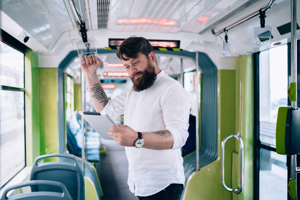 Bearded young man in white shirt and wristwatch standing in train and browsing social media in modern megapolis in soft daylight - Photo, Image
