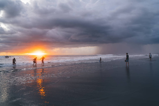 Group of people enjoying time on coastline with foamy waves splashing on sandy beach during dusk in cloudy evening in Bali - Φωτογραφία, εικόνα