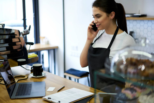 Professional brunette woman in apron having mobile phone conversation discussing business with delivery service operator - Fotoğraf, Görsel