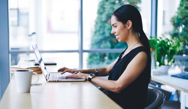 Side view of positive female remote worker with dark hair browsing netbook and smiling while sitting at table in contemporary workplace - Photo, Image