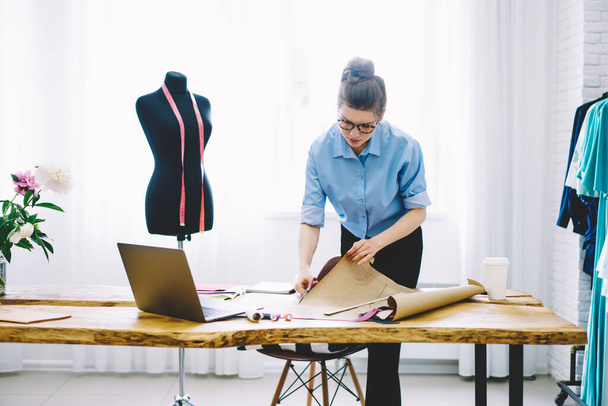 Young focused woman seamstress standing near table looking at craft papers with sketches of new clothes during workday in modern atelier - Foto, imagen
