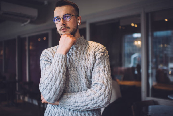 Pensive young bearded man in wireless headset and round eyeglasses standing with hand under chin and looking away with puzzled look on face - Φωτογραφία, εικόνα