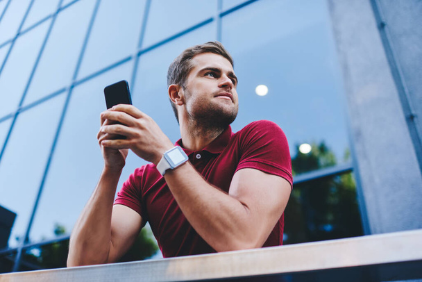 From below smiling young man in casual outfit and smartwatch browsing smartphone and looking away beside big modern building on blurred background - Zdjęcie, obraz