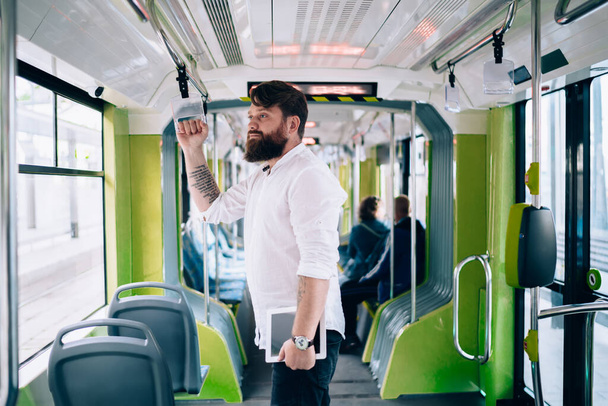 Side view of young bearded man in casual clothes with tablet and tattoo standing in train and looking away in soft daylight - Foto, Imagen