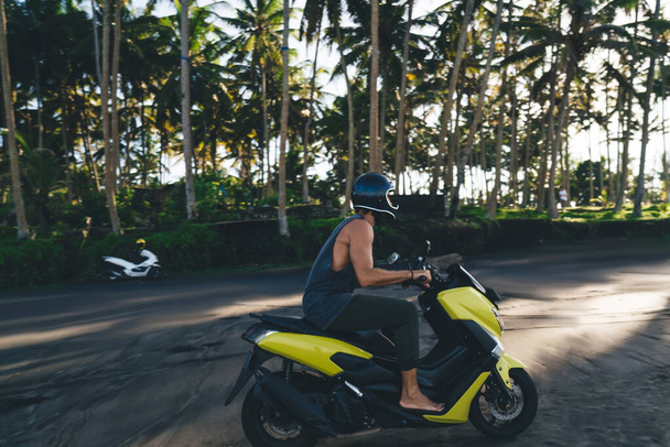 Side view of anonymous male biker wearing protective helmet and casual clothes driving motorbike along sandy beach near palm trees - Foto, Imagem