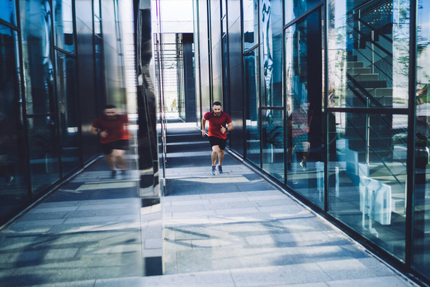 Full length of young athlete male in sportswear jogging by glass windows of modern office building on city street in sunny morning - Photo, image