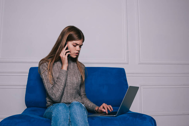Frowning female student in casual clothes calling on mobile phone and browsing on netbook while sitting on comfortable sofa in light room - Foto, immagini