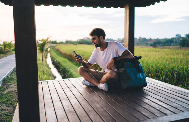Full body positive young male wearing casual summer clothes sitting with legs crossed in wooden alcove and browsing mobile phone in daylight - Fotografie, Obrázek