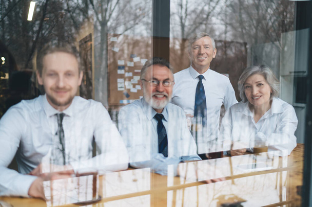 Through glass group of cheerful business partners in white formal shirts sitting at wooden table in office and looking at camera - Фото, зображення