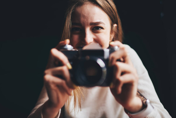 Young cheerful female photographer in white jumper taking photo with old fashioned digital photo camera looking at camera in dark room - Фото, зображення