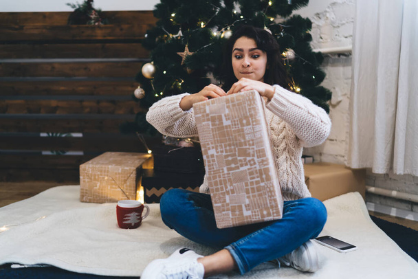 Wishful female unpacking box with gift while sitting near bright Christmas tree with garland and baubles in modern living room - Foto, imagen