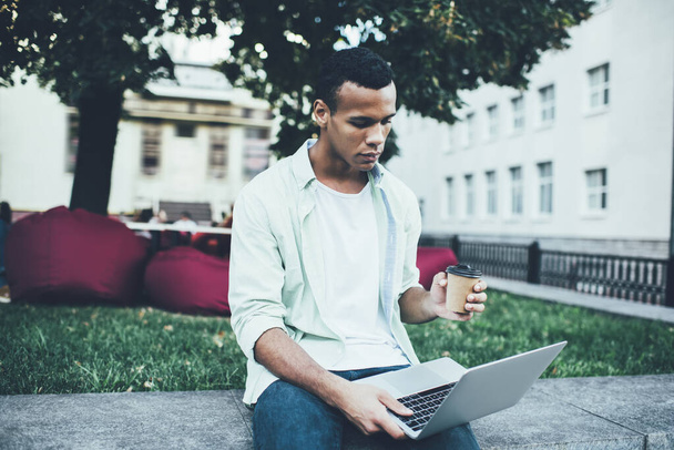 Serious young African American man surfing internet on netbook while having rest in city park and drinking hot beverage from disposable cup - Valokuva, kuva