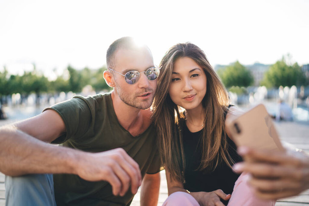 Positive multiethnic couple in casual outfit sitting on bench in sunny day in park and taking selfie together while resting - Valokuva, kuva