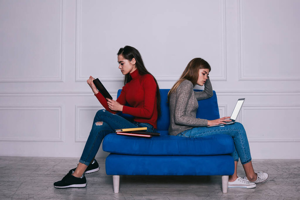 Side view of full body female students in casual wear sitting back to back on blue sofa and preparing homework with books and laptop - Foto, Imagem