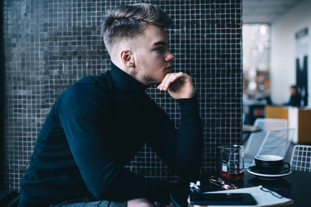 Side view of male employee with stylish haircut sitting at table with mug of hot drink and clipboard while thinking about future project - Fotoğraf, Görsel