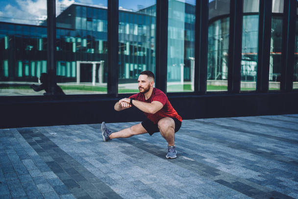 Full body of determined bearded male athlete doing exercises in city center and looking away while stretching legs and warming up - Foto, afbeelding