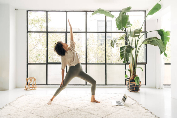Junge multiethnische Frau praktiziert Yoga zu Hause mit Online-Unterricht mit Laptop. Reverse Kriegerpose. Kopierraum. Gesunder und aktiver Lebensstil. - Foto, Bild