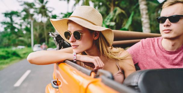 Pensive female tourist in hat and sunglasses leaning on hand and sitting in cabriolet near calm boyfriend while riding on road in summertime - Foto, immagini