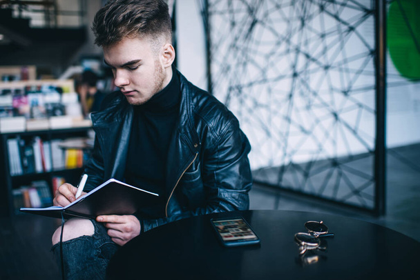 Crop serious young bearded male student wearing leather jacket sitting at table with sunglasses and smartphone and doing homework assignment in modern library - 写真・画像
