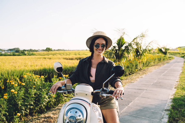 Portrait of joyful female tourist sitting on rented vintage moped smiling at camera while getting to travel destination in Vietnam - Zdjęcie, obraz