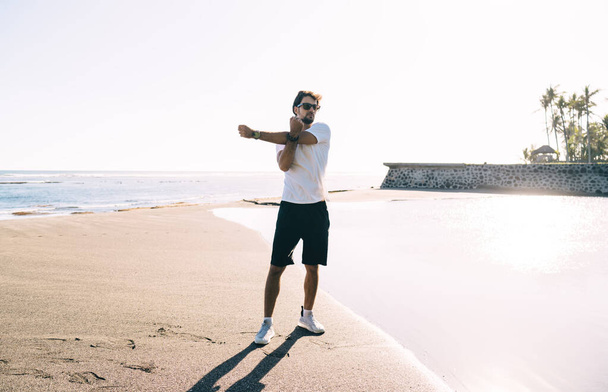 Full body of young slim man in sportswear and sunglasses stretching arm on beach while doing warm up exercises before workout - Φωτογραφία, εικόνα