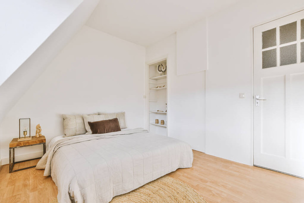 Pohodlná postel v blízkosti bílé zdi v podkrovní ložnici v moderním minimalistickém stylu s policemi a nočním stolkem - Fotografie, Obrázek