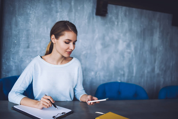 Young focused female freelancer sitting at table and taking notes while texting message and looking at screen of smartphone in office - Foto, Bild