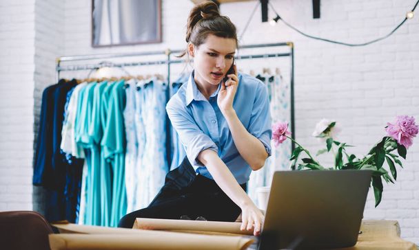 Low angle of serious businesswoman leaning on table and speaking on smartphone while browsing laptop and taking order of fabrics for work  - Photo, image