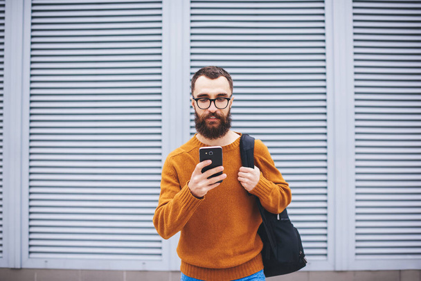 Serious bearded male in casual outfit with backpack looking at screen of smartphone while communicating with friends on street during rest - Фото, зображення