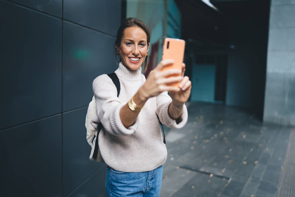 Pół długości portret wesoły kobieta turysta uśmiecha się do aparatu podczas korzystania z gadżet smartfon do klikania zdjęć - Zdjęcie, obraz