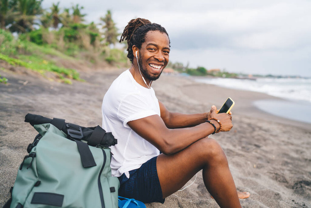Zijaanzicht van vrolijke Afro-Amerikaanse mannelijke reiziger in casual kleding met rugzak zittend aan zee en kijkend naar de camera terwijl hij geniet van muziek - Foto, afbeelding