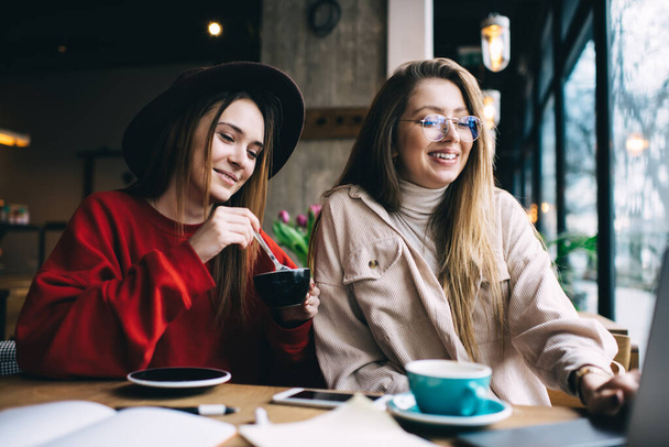 Smiling stylish girlfriends wearing casual outfits sitting and drinking hot coffee while studying together in contemporary cafeteria - Zdjęcie, obraz