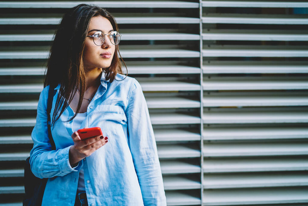 Contemplative hipster girl in classic eyewear holding smartphone device and thinking at urban settings - Zdjęcie, obraz