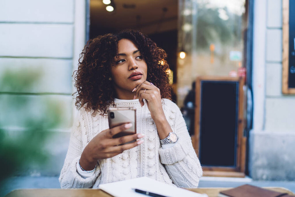 Pensive African American female in casual clothes sitting at wooden table with copybook and browsing mobile phone while resting in cafe - Foto, Bild