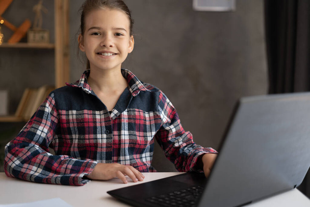 портрет счастливый ребенок смотрит в камеру, сидя за столом и с помощью ноутбука на дому, домашнее обучение концепции - Фото, изображение