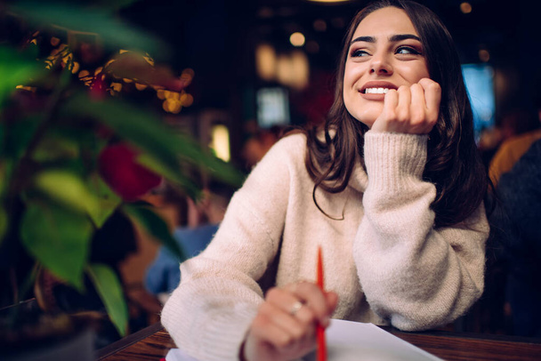 Cheerful young smiling lady in soft knitted sweater sitting at table with hand on chin and dreaming while looking away - Foto, immagini