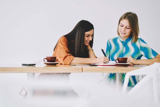 Happy female student taking note on notebook and sitting near female friend while working on homework together in light workspace against white wall - 写真・画像