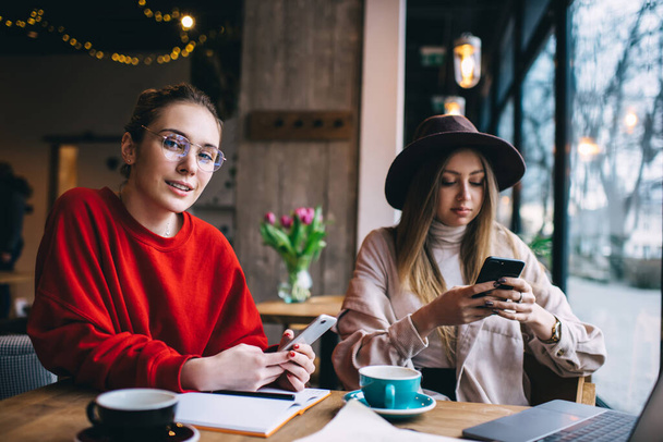 Young thoughtful female friends in casual clothes sitting at table with cup of hot drink and browsing web while spending time together in cafe  - 写真・画像