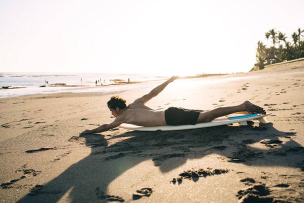 Side view full length sportive male surfer in shorts lying on surfboard and performing swimming moves while warming up before surfing on sea waves - Фото, зображення