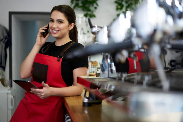 Cheerful brunette woman manage of coffee shor making ordering via mobile phone call standing at bar working space - Photo, Image