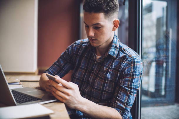 Concentrated male entrepreneur wearing casual clothes sitting at table and browsing smartphone while working on project in cafe with laptop - 写真・画像