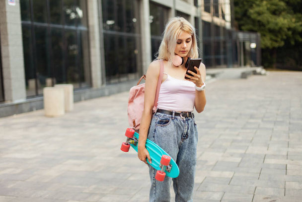 Cool young girl with skateboard using smartphone in the city - Foto, afbeelding